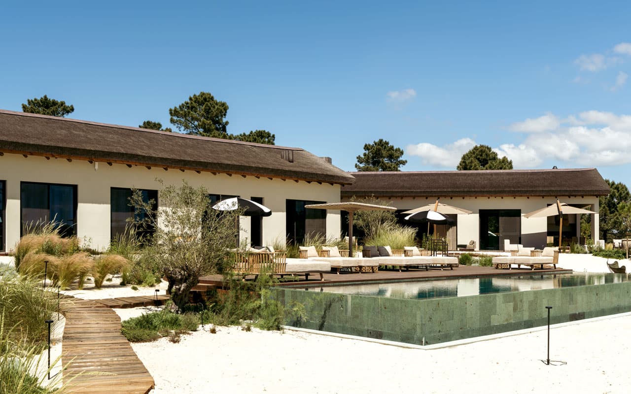 Luxury villa with Cape Reed thatched roof, timber decking, and infinity pool surrounded by natural sand landscaping in Comporta, Portugal
